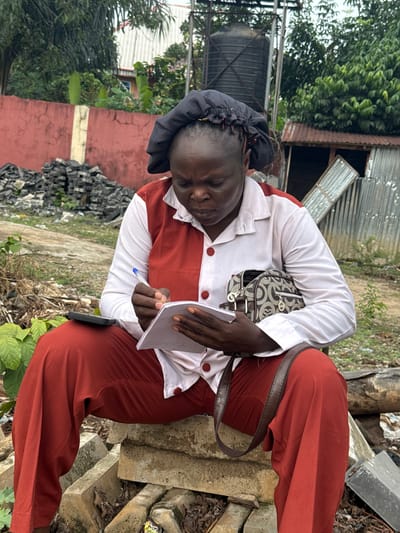 In Orifite, Nigeria, a woman wearing red pants, a white shirt with red trim, and a black head covering sits outdoors, hunched over a notebook and pen. She has a patterned handbag slung over her shoulder. She is positioned on what appears to be concrete blocks or debris. Behind her are scraps of metal and other debris. A water tank and buildings are visible in the background. There are also trees and plants.