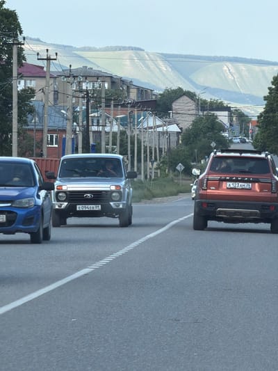 This media file captures a scenic view of a rural area in Russia, likely during the daytime given the bright natural light. The primary focus is on a road with vehicles, set against a backdrop of buildings and a wide, calm body of water, possibly a river or lake, reflecting the sky.
Several cars are visible on the road. Closest to the foreground is a blue car, followed by a silver off-road vehicle with the license plate "E094EB 09". Behind that, a red SUV is prominent, displaying the license plate "A 123AM 26". The road itself has a clear white line marking the lanes.
The setting appears to be a village or small town, with houses and utility poles lining the roadside. Trees are also present, adding to the natural environment. The water body in the background, bordered by a distant, tree-covered hillside, suggests a peaceful, perhaps somewhat remote, location. The overall impression is one of everyday travel through a picturesque Russian landscape. There are no discernible emotions or specific events taking place beyond the vehicles being driven on the road. The weather seems fair and clear, contributing to the pleasant scenery.