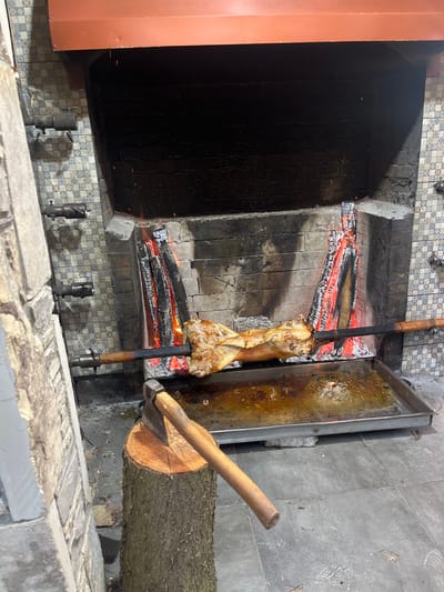 A piece of meat is being cooked over a fire in a large indoor grill. The meat, on a vertical wooden skewer, is positioned over hot coals within a brick and tile-lined structure. To the right of the grill, a metal tray is positioned to catch drippings, next to a large log with a partially embedded axe. The setting appears to be a kitchen or restaurant. The location in Miatli, Russia is provided as external information.