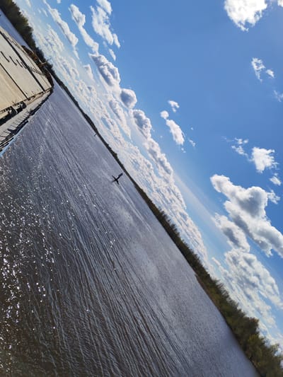 The image depicts a view from a pier over a body of water, presumably the Kama River, in Chaika, Perm Krai, Russia, given the provided location information. The water is textured with waves and reflects sunlight. Along the distant shore, a line of trees is visible. The sky is blue with scattered cumulus clouds. There is a floating navigational marker in the water. The pier's edge is visible on the left side of the frame. The image appears to be taken during the day.
