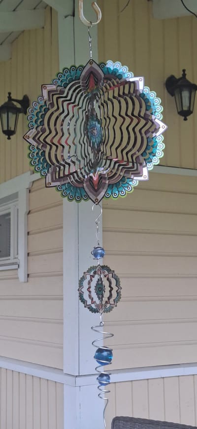 This media file shows a hanging wind spinner. The wind spinner is made of metal and has a colorful, mandala-like design with layered, wavy elements. It is suspended from a hook on an off-white, weathered surface, likely part of a porch or overhang. The background consists of light yellow, horizontally paneled siding, suggesting an outdoor setting.
The wind spinner is the primary object, and it appears to be stationary in the image. To the left of the main spinner, a black outdoor lantern is partially visible, mounted on the siding. Further to the right, the wind spinner extends with a thinner wire holding a smaller, similarly designed spinner, a blue glass bead, and a coiled section of wire with another blue glass bead.
The lighting suggests it is daytime, possibly overcast or in shade, as there are no strong shadows. The scene is tranquil, with no people or animals visible. The overall impression is one of outdoor decoration on a residential structure, possibly a porch or eaves. No visible text is present in the image. The location context of Orimattila, Finland, aligns with the outdoor setting and architectural style.