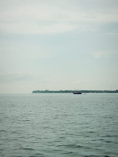The photograph captures a seascape with a hazy, overcast sky in Desa Pemenang Barat, Indonesia. The water appears calm with subtle ripples across the surface. A forested coastline stretches along the horizon. A dark-colored sailing ship with two masts is visible on the water, accompanied by a smaller boat nearby. A communications tower is also visible along the coastline.