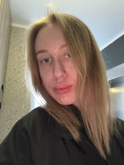 An image, rotated 90 degrees clockwise, captures a close-up portrait of a young woman. Her face is positioned towards the bottom-left of the frame, with her light brown, almost blonde hair cascading around her. She has striking blue or grey eyes, with the left eye clearly visible and gazing slightly upwards. Her lips are full and slightly parted, showing a natural pink hue, and she sports a subtle nose piercing on her left nostril. Her complexion is fair.

She is wearing a dark, likely black, garment, possibly a jacket or top, visible on the right side of the frame, with some texture visible in the fabric. The background is softly blurred but suggests an indoor setting. On the left, a light-colored wall or ceiling is visible, while the lower portion of the image reveals a speckled, light-toned surface, possibly a floor or countertop. Linear elements, potentially part of a building structure or ventilation, can be seen in the upper background. The lighting appears soft and even, highlighting her hair. There is no discernible specific activity other than posing for the photo, and her expression is neutral to slightly pensive. No text is visible in the image. The location context for this image is Kuleshovka, Russia.