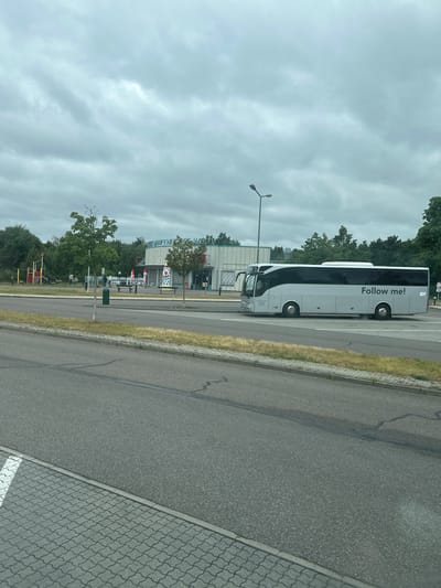 The image captures a grey, cloudy day in Schipkau, Germany. In the middle ground, a light grey tour bus with tinted windows is parked on the right side of a road. The bus has "Follow me!" written in large black letters on its side. To the left of the bus, a building with a curved glass facade is visible, featuring a sign that reads "BÖRWENEN WES". In front of the building, there are outdoor seating areas with red umbrellas, a bench, and a playground structure with red and yellow components. Several trees and greenery surround the scene. The foreground shows a paved road and a cobblestone sidewalk.