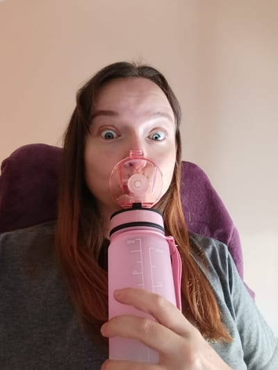 A woman with long brown hair and blue eyes is holding a pink water bottle in front of her face, with the bottle's lid positioned over her nose. The bottle has volume markings in ounces and milliliters. She is wearing a gray shirt and sitting in a chair with a purple cushion. The background is a plain beige wall. There is no indication of the location being in Águilas, Spain.