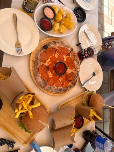 This media file captures a close-up, top-down view of a table laden with food and drinks, likely at an outdoor eatery given the dappled sunlight and paved ground visible in the background. The scene is set in Ptghni, Armenia.
The table features a variety of fast food items, suggesting a casual meal. Prominently displayed are two burgers, each with a skewer sticking out of the top bun, accompanied by servings of french fries in brown paper cones. A pepperoni pizza, cut into slices, is also a centerpiece. To the left, a plate holds several fried, breaded balls, possibly mozzarella bites or arancini, served with small dishes of pink sauce and ketchup.
Beverages include a bottle of what appears to be a dark-colored soda or juice with a "MJ MAX&JACK'S" label featuring a cherry graphic and the text "ALMOND". Another bottle, mostly obscured by the frame, is blue and white.
Several white plates and cutlery, including a fork, are arranged on the table, indicating that diners are present or about to begin their meal. An ashtray is also visible. Some of the food items are served on wooden trays. The overall impression is one of a shared meal with friends or family, enjoying a sunny day. The lighting suggests it's daytime, possibly late morning or afternoon. The visible text on the bottles and some packaging includes "MAX&JACK'S", "ALMOND", "MJ", and "DARKSIDE".