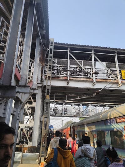 The image displays a busy railway station platform in Bardhaman, India. A multi-level overhead pedestrian footbridge, constructed from grey metal girders and lattice-patterned railings, spans across the scene, featuring a corrugated roof structure. A yellow rectangular sign marked "106 20B" is affixed to a vertical support beam of the footbridge on the left side. Overhead electrical wires and insulators are visible, typical of an electrified railway environment, running below the footbridge and across the tracks.
On the right side of the frame, a blue and cream-colored passenger train, identified by Hindi script on its side (e.g., "हमसफर एक्सप्रेस" - Humsafar Express), is stationary at the platform. Further down the tracks on the left, a red-colored train is partially visible.
The platform is densely populated with individuals, primarily seen from behind, walking from the left foreground towards the right midground. These individuals are dressed in various casual attire and many are carrying luggage, including backpacks and tote bags. On the extreme left foreground, the partial profile of a male subject with dark hair and a beard/mustache is visible, looking towards the right. Several other figures are discernible traversing the lower level of the overhead footbridge in the background. The sky above the structures is clear and light blue.