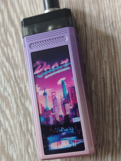 A purple and pink pod-style electronic cigarette is shown lying on a light-colored, wood-grain surface. The electronic cigarette has a black mouthpiece and a panel on the front displaying a vibrant, neon-infused image of a city skyline at night. The city appears to be a stylized representation, possibly inspired by cyberpunk aesthetics, with tall buildings, illuminated signs, and vehicles on the street. The text "Dinao" is visible in a retro font above the city scene, and "Pasito II" is in the lower portion of the panel. The overall impression of the city graphic is futuristic and retro. The electronic cigarette is positioned diagonally across the frame. The background is a wooden surface with a prominent grain pattern. There is no indication of the city of Лычково, Russia, in the image.