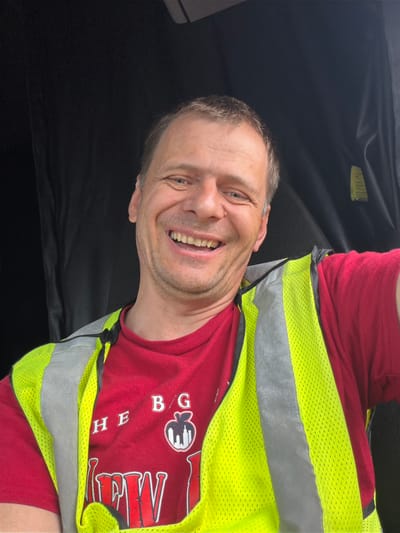 The media file is a portrait-oriented photograph of a man smiling and looking towards the camera. He is wearing a red t-shirt and a bright yellow reflective vest with gray reflective stripes. The man has short, light brown hair and stubble. He appears to be in a vehicle, indicated by the dark interior visible behind him, which is likely the headliner and a black seat or surface. The lighting suggests it is daytime. The man's expression conveys happiness or amusement. There is some text visible on his t-shirt, which appears to be a logo and some lettering. The logo is a stylized apple with a building inside, and the text reads "THE BIG APPLE." The photograph is taken at an angle, making it appear slightly tilted. The setting is likely Chattanooga, United States of America, as provided in the context. No specific weather or time of day cues are evident beyond it being daytime.