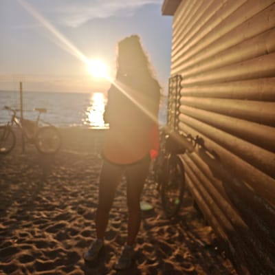 A person stands on a sandy beach with a wooden building to their right, facing the sea during sunset. The sun, low in the sky and casting a bright glare, is reflected on the water. A bicycle is parked to the left of the person, and another bicycle is near the wooden building. The ground is covered in sand with footprints and some debris. The wooden building has horizontal planks and appears to be a small structure like a shed or kiosk. The overall scene suggests a recreational setting by the sea at dusk. The location is identified as Ленинское сельское поселение, Ukraine.