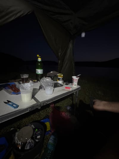 The image depicts a campsite at night, with a tent visible in the foreground and a table laden with items. The scene is dimly lit, suggesting it's either very early morning or late at night. The presence of a tent, along with what appears to be fishing gear in a bag, indicates an outdoor camping or fishing trip.
On the table, several objects are scattered: two plastic cups, one filled with a white liquid, the other with a clear liquid and some condensation; a green glass bottle with a yellow cap, and another bottle behind it. There's also a red lighter, a small white object possibly a pill container or dispenser, and some food items wrapped in plastic. A blue handled multi-tool, resembling scissors or a small knife, lies on the table. In the background, a person's legs are visible, with one foot wearing a dark sandal or flip-flop, suggesting a casual, relaxed setting. The ground around the campsite is covered in grass.
To the right of the table, a black bag is open, revealing spools of fishing line and other fishing accessories. A plastic water bottle with some text in Cyrillic script is also on the grass near the bag. The overall impression is of a campsite that has been set up for an overnight stay, possibly for fishing, with items laid out for use. The lack of distinct weather cues suggests it's likely a calm night. The location context of Debelets, Bulgaria, further supports the idea of an outdoor activity in a rural or natural setting.