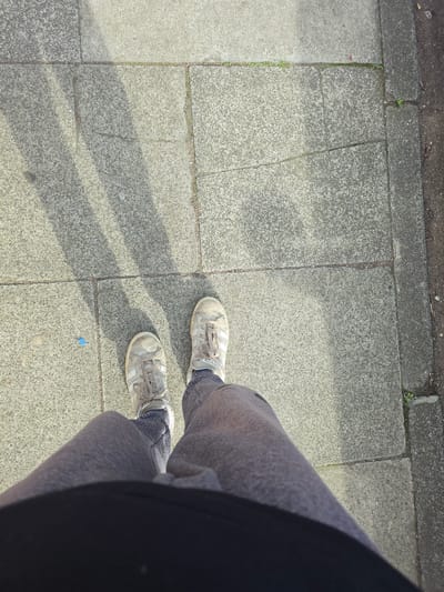 The image shows a pair of feet wearing light-colored sneakers and grey sweatpants, standing on a paved sidewalk. The sneakers appear slightly worn and dirty, with white laces and the signature three stripes of Adidas visible. The sweatpants are a textured grey fabric. The subject is standing on a section of concrete paving slabs, some of which are cracked and uneven. Sunlight casts distinct shadows of the person's legs and feet onto the pavement, indicating it is daytime. A small, bright blue object, possibly a piece of plastic or a cap, is visible on the ground near the person's right foot, along with what looks like a coin. The edge of a dark fabric, likely clothing such as a jacket or shirt, is visible on the right side of the frame, obscuring part of the subject's body. The background suggests an outdoor urban environment with the concrete slabs continuing into the distance. The scene is brightly lit, suggesting it is a sunny day, possibly in the late morning or early afternoon. No text is visible in the image. The overall impression is a candid snapshot of someone standing outdoors on a sunny day.