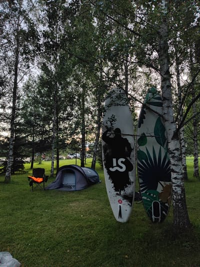The image depicts a serene camping scene in the city of Saltanovo, Russia. Two paddleboards, one with a silhouette design and the other adorned with tropical leaves, lean against birch trees. In the foreground, a dark grey tent is set up on a lush green lawn, with a foldable orange life jacket draped over a black camping chair beside it. The background reveals a cluster of birch trees and a hint of a distant structure, suggesting a tranquil, natural setting on the outskirts of the city. The lighting indicates it's either early morning or late evening, with the sky displaying subtle hues of orange and pink.