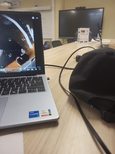 The image shows a laptop computer with a black screen showing a video playing. The laptop is open on a wooden table in a room, along with a black cap sitting next to it. The laptop is connected to a black cable, possibly a power cord, and there is a black cord running across the table. On the table next to the laptop is a white piece of paper with text that is not readable in the image. There is a large, black flat-screen monitor behind the laptop. The monitor is turned off, with a black screen, and there is a small white webcam in the top center of the screen. The room is sparsely furnished with a few chairs and a whiteboard with writing on it. It appears to be a meeting room. The coordinates provided place this image in a building in Calais, France.