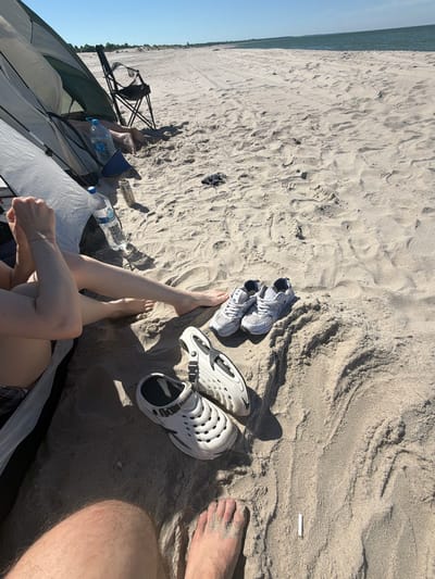 A sunny day at the beach in Povarovka, Russia, finds two pairs of shoes, one white clogs and one pair of athletic shoes, placed on the sand near a tent. The scene is likely a campsite as a portable chair is set up nearby, and two water bottles and a can are visible. A person's legs are visible in the foreground, resting by the tent, and another person's feet are also in the frame. The background shows a vast expanse of sand leading to the calm blue sea. The overall impression is one of leisure and relaxation on a warm day by the coast.