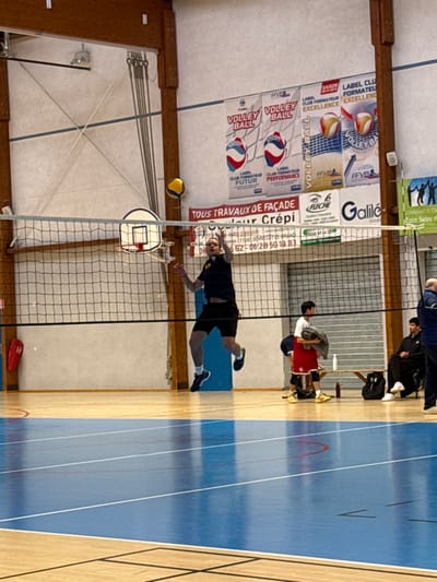 This image depicts an active indoor volleyball match in a gymnasium located in Sennecey-le-Grand, France, as indicated by banners displaying the postal code "71240 SENNECEY LE GRAND." The primary subject, a male athlete, is captured mid-air above the blue-marked wooden court, positioned near the volleyball net. He wears a dark short-sleeved shirt with a yellow logo, black shorts, and black athletic shoes, with visible tattoos on his left forearm. His arms are raised, suggesting an attempt to spike or block a yellow and blue volleyball, which is airborne slightly to his upper left.
The gymnasium features light-colored walls with vertical wooden structural beams. Numerous banners are displayed on the far wall, including those for "VOLLEY BALL" from the French Volleyball Federation (FFVB) for the "Saison 2018-2019" and advertisements for local businesses such as "TOUS TRAVAUX DE FAÇADE Couleur Crépi," "Fleche," and "Galilée Group." A basketball hoop with a white backboard is visible behind the volleyball net on the left.
On the right side of the background, beyond the volleyball net, three male individuals are present near a wooden bench: one stands, dressed in a white shirt and red shorts, holding a grey item; two others are seated on the bench, wearing dark attire. A fourth male is partially visible on the far right. A white water bottle and a black backpack are on the floor near the bench. A retractable metal gate or shutter is visible on the back wall, and a red rescue buoy-like object hangs on the far left wall.
