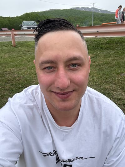 A man in a white t-shirt smiles at the camera in a selfie. He has short, dark hair that is slicked back and his skin is slightly red. In the background, there is a grassy area, a guardrail, trees, and a mountain range. The man appears to be outdoors, possibly in a park or rural area. The image does not contain any discernible landmarks or specific details that would place it within the city of Malka, Russia.