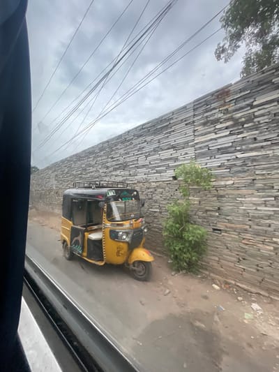 The image shows a yellow auto-rickshaw parked on a dirt road next to a stacked stone wall. The sky is overcast with visible power lines. A small green bush grows in front of the wall. The auto-rickshaw has a black roof and is partially open. The scene appears to be in Bethamcherla, India.