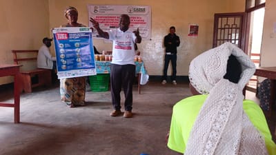 In Kamakombe, Democratic Republic of the Congo, a man in a white t-shirt and dark pants gestures animatedly while standing behind a table covered with a patterned cloth, on which several jars are arranged. He is addressing a group of people. To his left, a woman in a headscarf and patterned dress holds a large poster with the text "STOP EBOLA" and other French writing. To the right of the man, another man in a black hoodie and dark pants stands with his arms crossed, observing the scene. In the foreground, a person wearing a bright green shirt and a white crocheted headscarf is partially visible from the back. The environment appears to be an indoor space with concrete flooring, yellow-painted walls, and a wooden door in the background. A partially visible wooden bench is present on the left.