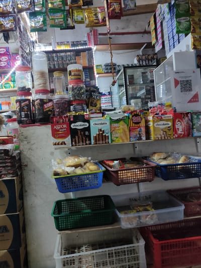The image depicts the interior of a small convenience store in Meunasahbaet, Indonesia. Shelves are stocked with a variety of packaged snacks, candies, cookies, and drinks. Jars of bulk items like candies and snacks sit atop the shelves. Several plastic baskets of pastries or bread are visible on lower shelves. Packaging for brands like Kopiko, Astor, and Chocolatos are visible. An electronic payment QR code is affixed to the storefront. The scene suggests a small, local business catering to immediate consumer needs.