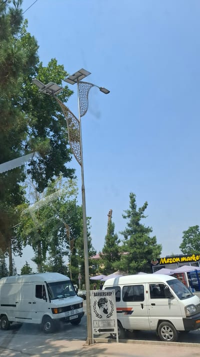 This media file depicts an outdoor scene with a clear blue sky. In the foreground, a decorative street light with solar panels is visible against the sky and trees. To the right, a white van is parked on the side of the road, and a sign for "BARBERSHOP" with an illustration of a man's head and the text "ERKAKLAR SALONI" is propped up nearby. Another white van is partially visible in front of the parked van. Trees and buildings are present in the background, suggesting an urban or suburban setting. The orientation of the image is rotated, making it appear sideways. The lighting suggests it is daytime.