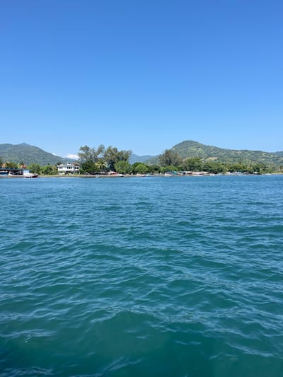 The image captures a coastline in Pemenang Timur, Indonesia. On the left side, lush green mountains and trees form the landscape, interspersed with houses and other structures that define the coastal settlement. The right side of the image presents a vast expanse of blue ocean water with gentle ripples.