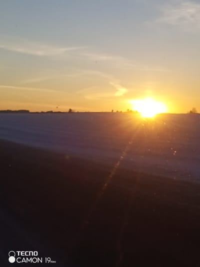 The image captures an outdoor, snow-covered landscape at sunrise or sunset in Bal'zyashur, Russia. The horizon line bisects the image horizontally, separating a gradient sky from the ground. The sun, a brilliant, intense light source with visible rays and lens flare, is positioned near the center-right of the horizon. The sky directly around the sun is colored in vibrant yellows and oranges, transitioning to a pale blue in the upper portion, where sparse, wispy clouds are present. The ground appears as a vast, flat expanse of white, indicative of snow cover. Along the distant horizon, indistinct dark silhouettes, consistent with trees or structures, are barely discernible. The immediate foreground is a dark, undefined surface, possibly an interior car dashboard, through which the scene is viewed, displaying subtle light reflections from the sun. No individuals, specific actions, or interactions are visible. A watermark reading "TECNO CAMON 19 Neo" is present in the lower-left corner.