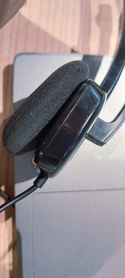 The image shows a close-up, overhead view of a black headset resting on a light gray surface, likely a laptop. The headset has a foam earpad and a glossy black casing. A black cable extends from the headset towards the bottom left of the frame. The background consists of a wooden surface with soft lighting casting shadows. The scene is static and does not depict any specific location within Pagedangan, Indonesia, as there are no identifying landmarks or contextual clues. The focus is solely on the headset and its immediate surroundings.