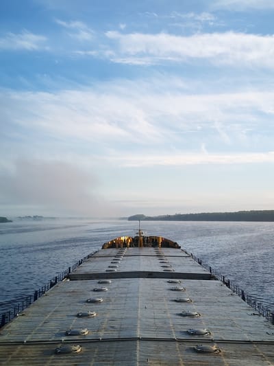 A long, gray, flat-topped vessel dominates the foreground, stretching toward the horizon. Circular hatches punctuate its surface in a symmetrical pattern. A low, blue railing runs along the sides of the vessel. In the distance, land with trees lines both sides of the water. Fog or mist obscures portions of the landscape. The sky above is light blue with wispy clouds. No people are visible. The location is not necessarily in Anisimovo, Russia.