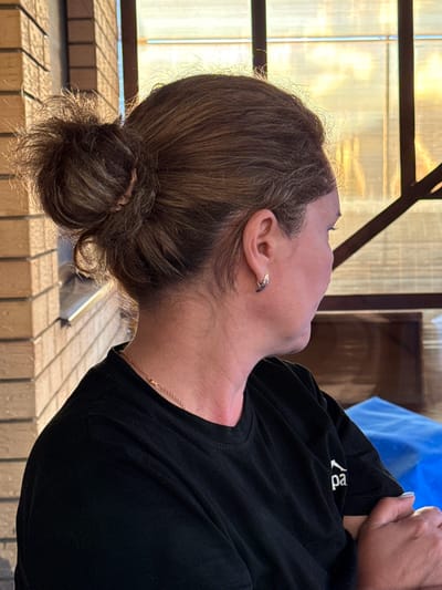 This is a close-up, rotated image of a person's head and upper torso. The person has brown hair pulled back into a bun at the nape of their neck, with some wisps of hair around their forehead and ears. They are wearing a black t-shirt with a white logo on the left side of the chest, which appears to read "ropa". A thin, delicate gold chain necklace is visible around their neck. The person has an earring in their left ear. The background suggests an outdoor setting, possibly a patio or balcony, with wooden structures and a translucent, yellowish material that could be a screen or awning. The lighting suggests it is daytime, with sunlight illuminating the scene and casting shadows. The overall impression is a candid, informal portrait.