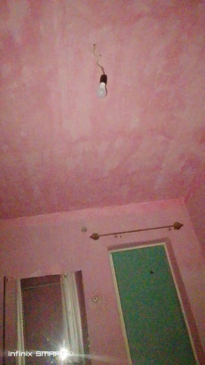 The image shows a pink ceiling with a bare light bulb hanging from a wire. There is a mirror on the left side of the image reflecting a bright light. On the right side of the image, there is a green door with a white frame. A curtain rod with decorative elements is mounted above the door. The bottom left corner of the image shows a watermark that reads "Infinix SMART 10". The scene appears to be indoors, likely a room in a house. The lighting is dim, with the primary light source being the exposed bulb.