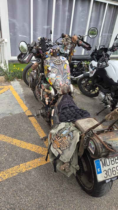 The image depicts a heavily customized "rat bike" motorcycle parked on asphalt with yellow lines. The bike has numerous modifications and attachments, including stickers, chains, bags, and miscellaneous objects. Its license plate reads "MUBB 3066." Another motorcycle, a white BMW, is visible behind the rat bike. The location appears to be outdoors, near a building with glass windows. The city is Altura, Spain.