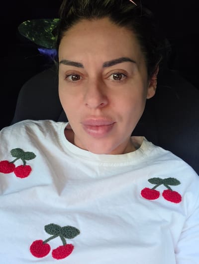 In Ravnogor, Bulgaria, a woman with brown eyes and dark hair pulled back sits in a car, looking at the camera. She is wearing a white t-shirt adorned with embroidered red cherries. The background is dark, with a hint of greenery visible through a window.