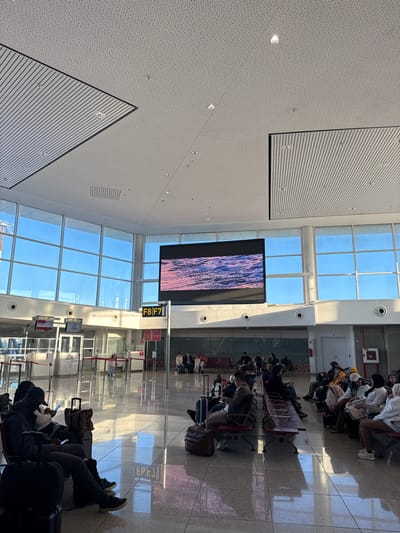 The image depicts the interior of a modern airport terminal, specifically a departure or waiting area, located in Nouaceur, Morocco. The scene is brightly illuminated, with strong natural light entering through large, multi-paneled floor-to-ceiling windows that reveal a clear blue sky outside. The ceiling is high, featuring a combination of white perforated acoustic panels and slatted metallic panels, with numerous recessed lights.

The floor is a highly polished, light-colored tile, exhibiting significant reflections of the ceiling, windows, and individuals present. In the mid-ground, a large digital display screen is centrally positioned, exhibiting an image of water ripples with text partially visible, including "A NEW WAVE OF LIVING." Below and to the left of this screen, a yellow gate sign clearly indicates "F8 F7."

To the left, beyond the main waiting area, are gate entrances, labeled "GATE F8" and "GATE F7," with red retractable stanchions and barriers in place. Several individuals are visible in this area, including one person near the gate entrance.

The foreground and mid-ground are populated by approximately 20-30 individuals, predominantly seated on rows of connected, dark-colored waiting chairs with metal frames. Most individuals are dressed in casual attire. Many are observed looking down at mobile phones or other personal devices. Various pieces of luggage, including suitcases and bags, are placed on the floor next to or in front of the seated passengers. Reflections of the seated individuals and their luggage are clearly visible on the polished floor. No active boarding or disembarking is depicted; the scene conveys a state of waiting. A red fire extinguisher cabinet is visible on a pillar on the far right.