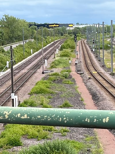The image depicts a view of a railway line in Dudelange, Luxembourg. In the background, a large building with a prominent "GOODYEAR" sign is visible, suggesting proximity to industrial or commercial facilities. The foreground is dominated by railway tracks, overhead power lines supported by numerous poles, and overgrown vegetation along the sides of the tracks. The sky is overcast, indicating a cloudy day. The scene is devoid of any people or vehicles, presenting a static view of the infrastructure. A large green pipe with white speckles runs horizontally across the lower part of the image, partially obscuring the railway tracks and surrounding landscape.