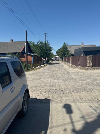 A clear, bright day in Sarny, Ukraine, is captured from a slightly elevated, rotated perspective. The scene looks down a dusty, gravel-lined road bordered by houses and fences on the left and a paved sidewalk on the right. A light-colored, modern SUV is parked close to the camera, its side and spare tire visible.
The road is flanked by a mix of buildings. On the left, closer to the camera, are houses with corrugated metal roofs and wooden siding, interspersed with trees. Further down the road, more houses and fences are visible, along with utility poles and power lines stretching across the bright blue sky. On the right side of the road, the sidewalk is made of interlocking grey bricks. A distinct shadow of what appears to be a lamppost or similar structure falls across the paved surface.
The overall impression is of a sunny, ordinary day in a residential area. There are no people visible, and the focus is on the environment and the parked vehicle. The quality of light suggests it is likely midday.