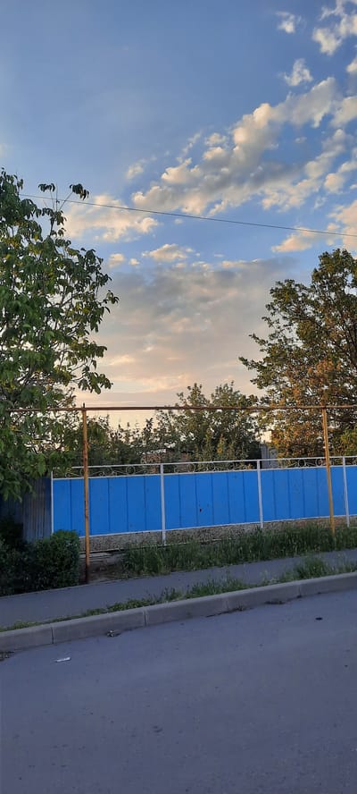 In the city of Shakhty City District, Russia, a scene unfolds with a blue fence, its metallic structure supporting decorative elements at the top. The fence is bordered by green foliage and grass, hinting at a residential or garden area. The sky above is a canvas of blue, adorned with scattered clouds, some of which are catching the warm hues of what appears to be either dawn or dusk. Trees with vibrant green leaves stand on either side of the fence, framing the view and adding to the natural ambiance. A paved road with a curb runs in the foreground, suggesting a street or pathway adjacent to the fenced area. The overall atmosphere is tranquil, with the natural elements blending seamlessly with the man-made structures.