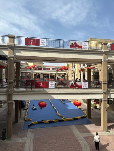 A multi-story building with balconies is decorated with red banners and fish-shaped lanterns. A blue, river-like structure with yellow and black borders is set up on the ground. Red lanterns are placed on it. People walk by. The sky is visible above the building.