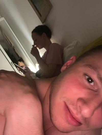The image, rotated 90 degrees counter-clockwise from its likely original orientation, features a close-up selfie taken indoors, contextualized to be in Vitry-sur-Seine, France. The primary subject is a young man whose face occupies the majority of the foreground, specifically the right side of his face from chin to forehead. He looks directly at the camera with dark brown eyes and a neutral to slightly smiling expression. His fair skin shows some flushing on the cheeks and light stubble on his chin and upper lip. Part of his bare shoulder or upper arm is visible in the lower right, suggesting he is shirtless or wearing minimal upper body attire.
In the background, a reflection in a mirror or polished surface reveals another person with dark hair, wearing a light-colored (possibly pink) short-sleeved top. This individual appears to be holding a yellow or orange item to their mouth, as if eating. A bright artificial light source is visible in the reflection, positioned between the two figures. The reflection also shows a white surface, likely a table or dresser, and a framed picture on the wall to the left of the reflected person. The overall dimness with artificial lighting suggests an evening or nighttime setting within a domestic space, likely a bedroom. No discernible text is visible in the image.
