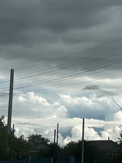 The image depicts an overcast sky over a rural landscape. Several power lines crisscross the frame, supported by utility poles. In the foreground, the tops of trees and parts of buildings are visible, suggesting a residential area. The sky is filled with dark, heavy clouds, indicating a potential for rain or stormy weather. The overall impression is of a somber, atmospheric scene. The location is identified as Тимирязев ауылдық округі, Kazakhstan.