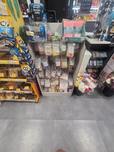 This is a medium-angle, full shot of a retail display area in a store, likely a convenience store or supermarket. The scene is a close-up of shelves stocked with various food items and a few non-food items.
Prominently featured are several packages of "KP Original Salted Peanuts" and "KP Salt & Vinegar Peanuts" in blue and yellow packaging. Below these, a yellow wire display rack holds containers of nuts and dried fruits, including pistachios and almonds, with visible price tags like "£3.95" and "£4.29". Also visible on this rack are boxes labeled "KP Crackers" and what appear to be biscuits.
To the left, there are several blue boxes of "VIDAL Ocean Jelly" with cartoon octopus and fish characters. Further left, a section of a shelf displays sunglasses on a rack. A price board nearby shows "2 for £4.50", "5 for £9.95", and "7 for £12.95".
On a higher shelf, there are multiple packets of sweets and candies, including those labeled "Kavanaghs" with flavors like "Sherbet Lemon", "Choco-late", and "Fruit". There are also boxes of "Halls" and "Locket" lozenges. In the center, a large bouquet of mixed flowers in shades of pink, white, yellow, and purple is displayed in a black pot. Below the flowers, more packets of sweets are visible, some with labels like "White Mice" and "Peach Ring".
The background consists of a gray tiled floor and the continuation of retail shelving. The lighting appears to be overhead store lighting, suggesting it is daytime. The overall scene conveys a typical retail environment focused on impulse purchases and snacks. No people are visible in the image.