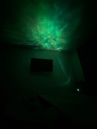 The image depicts a dimly lit room, likely a bedroom, with a projector casting a green, nebula-like light pattern onto the ceiling and upper walls. A black, rectangular television screen is mounted on the left wall. In the lower right corner, a faint glow suggests a small light source, possibly a lamp, on a dark surface. The rest of the room is shrouded in shadow, with indistinct shapes suggesting furniture or bedding. The scene appears to be indoors, with no visible windows or outdoor elements. The location in Zhigulyovsk, Russia, cannot be determined from the visual information provided.