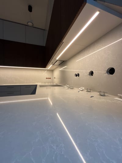 The image shows a modern kitchen under construction or renovation in Tartu, Estonia. The scene is captured from a low angle, emphasizing the countertops and cabinetry. A light gray, marbled countertop with embedded linear LED lighting extends across the foreground and midground. Behind the countertop, dark gray cabinets with minimalist drawer pulls are visible. The backsplash is a light-colored stone material with veining, also featuring integrated linear LED lighting.
On the backsplash, there are several circular cutouts, some with electrical wiring exposed, suggesting unfinished electrical work. There are also some small metallic fixtures and white electrical components scattered on the countertop. A black induction cooktop is set into the countertop. Above the cooktop, a modern range hood is installed against the wall. The upper part of the image reveals dark wood paneling on the ceiling and a dark vertical accent wall. A single pendant light fixture is suspended from the ceiling. The overall impression is one of a contemporary kitchen design with a focus on integrated lighting and sleek finishes.