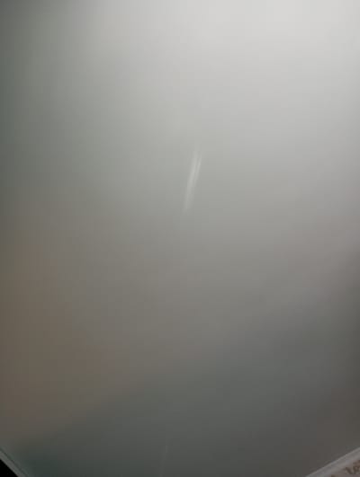 The image displays a uniform, light gray interior surface, likely a wall or ceiling, located within the city of Яромаска, Russia. A distinct, elongated, bright white vertical streak is centrally positioned in the upper-right quadrant of the frame, approximately 10-15% from the right edge and extending downwards. The surface exhibits subtle variations in tone, with diffuse lighting originating from an unseen source, causing a slight gradient from lighter values in the upper-right to darker values in the lower-left. In the extreme bottom-right corner, a partial view of a patterned, light-colored edge or surface element is visible, possibly indicating a floor or a baseboard. No individuals, additional specific objects, or discernible actions or interactions are present within the frame.