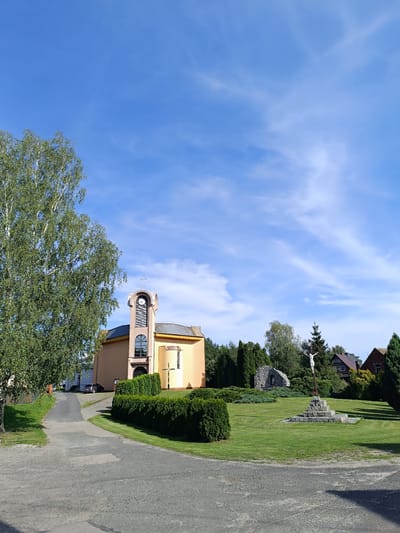 The image depicts a clear day in Czekanów, Poland, with a bright blue sky dotted with wispy clouds. In the foreground, a gravel path leads towards a modern church with a peach-colored facade and a prominent clock tower topped with a cross. To the left of the path, a large birch tree with lush green foliage stands tall. In front of the church, a manicured hedge lines a grassy area. To the right, a stone monument with a crucifixion scene featuring a white Christ figure on a cross is visible. Behind the monument, a cluster of trees and a small stone structure can be seen. Further back, residential buildings are nestled amongst the greenery. A dark-colored car is parked on a road leading to the church, and another building is partially visible behind the main church structure. The scene is serene and evokes a sense of rural tranquility.