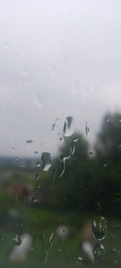 The image is a close-up, vertical shot of raindrops on a windowpane. The raindrops are varied in size and shape, with some appearing as small droplets and others as larger, elongated streaks, indicating they are running down the glass. The background is blurred, suggesting an outdoor scene viewed through the window. The blurred background shows a hint of green foliage, possibly trees and grass, and a muted sky that appears overcast or hazy. There are no people, specific objects, or actions clearly discernible. The overall impression is that of a rainy day viewed from indoors. The location is not determinable from the image itself, therefore it cannot be specified to be in Kudymkar, Russia.