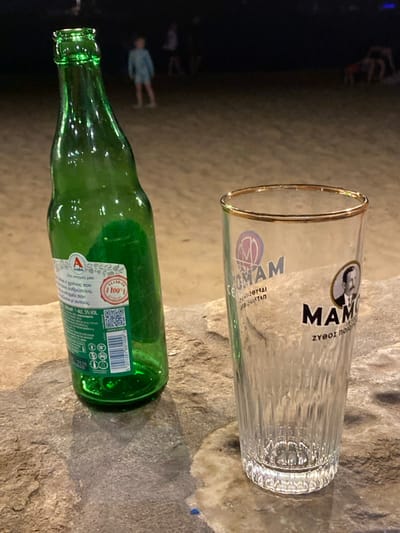 A nighttime scene depicts an empty green glass bottle and an empty clear glass with a gold rim, positioned on a light-colored, textured stone surface in the foreground. The green bottle, labeled "Alpha" beer with "ALC. 5% VOL," stands slightly tilted. The clear glass is branded with "MAMOS" and a portrait, featuring vertical ridges on its lower half. In the blurred background, a sandy beach extends into darkness, where several indistinct human figures are visible; one figure, appearing to be a child in light-colored attire, stands centrally in the mid-distance. This scene is situated on a beach in the city of Φάρος, Greece.
