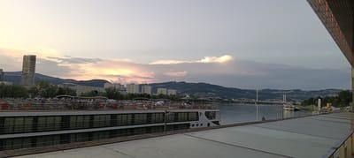 In Linz, Austria, a wide shot captures a twilight scene by a river. In the foreground, the upper deck of a cruise ship is visible, with tables, chairs, umbrellas, and people seated and standing. The ship has multiple levels of windows. To the left, a modern high-rise building stands against a backdrop of rolling hills and other urban structures. Across the river, more buildings and a suspension bridge are visible in the distance, with hills forming the horizon. The sky is filled with dramatic clouds illuminated by the setting sun, casting warm hues. The right side of the image shows the edge of a contemporary building with illuminated vertical strips.