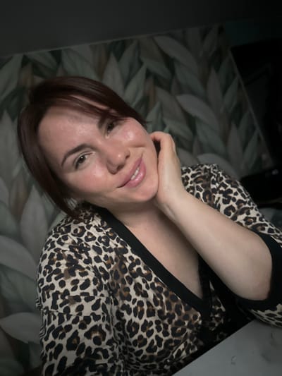An indoor portrait features an adult woman looking directly at the viewer with a pleasant, gentle smile, showing a hint of her teeth. She has dark brown hair, cut to approximately chin length, framing her face. She is wearing a top or dress with a distinct brown, black, and white leopard print pattern, accented with solid black trim around the neckline. Her left arm is bent, and her hand rests gracefully near her chin and neck.
The background consists of a dark, subtly textured surface, likely a wall or fabric, adorned with a repeating pattern of stylized, muted grey-green leaf shapes. To the far left and top right, darker, less defined areas are visible, possibly indicating furniture or other parts of the room. The lighting is soft and even, illuminating her face and upper body, suggesting an indoor setting without strong natural light cues for time of day. Her expression conveys contentment and friendliness. No text is visible within the image. The provided location context is Черёмушки, Russia.