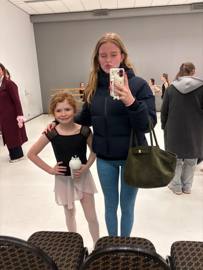 A woman in a black puffer jacket and blue leggings takes a selfie with a young girl in a black leotard and pink tutu. The girl holds a white water bottle. They appear to be in a dance studio with a barre and other dancers visible in the background. The woman is holding a large green tote bag. Rows of chairs are also visible.
